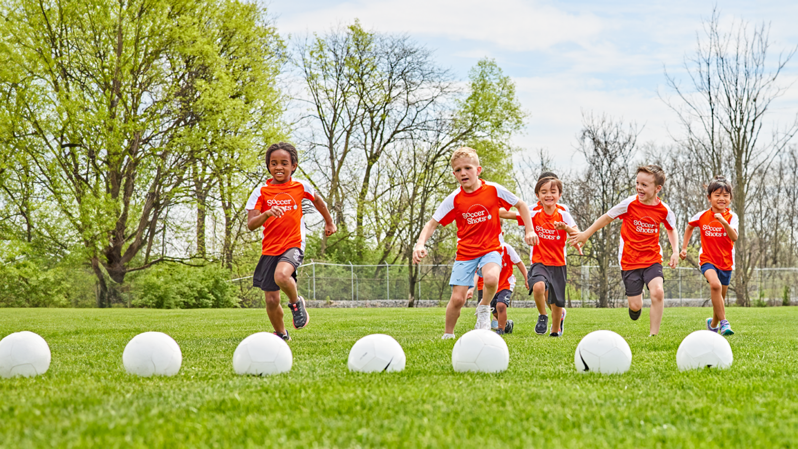 Row of kids in Soccer Shots jerseys running towards a line of soccer balls.